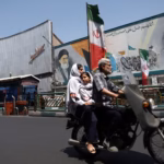 Orang-orang berkendara melewati mural yang menggambarkan mendiang pemimpin tertinggi, Ayatollah Ruhollah Khomeini, di Teheran/ Foto: The Guardian
